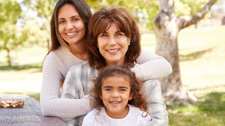 Three generations of women seated outside at a picnic table are smiling at camera.