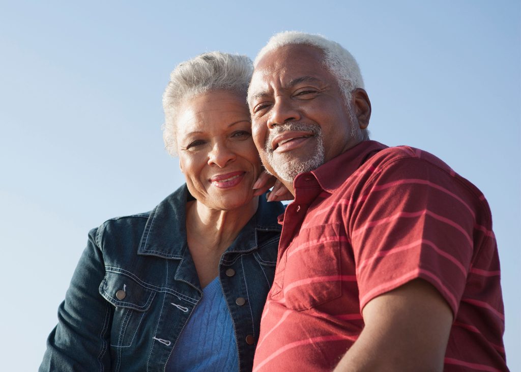 Mature couple outdoors in sunlight holding each other close and smiling at the camera.