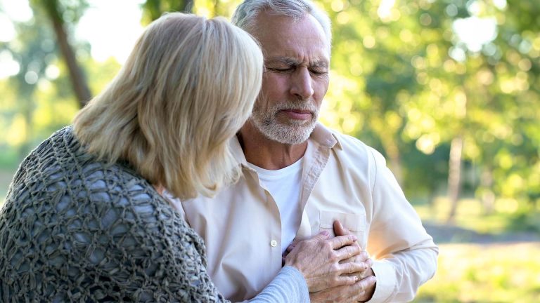 Old couple sitting in park and the man is clutching his chest.