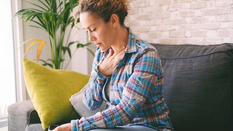 A woman sitting on a couch at home, looking distressed with her hand on her chest, possibly experiencing discomfort.