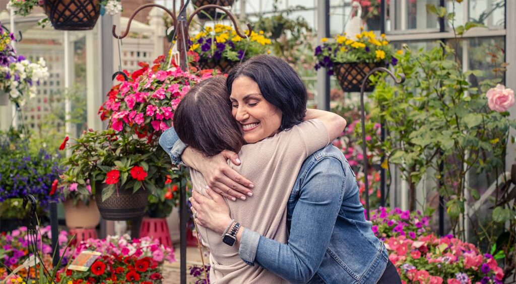 Alexis and woman friend embracing in a flower-filled greenhouse.