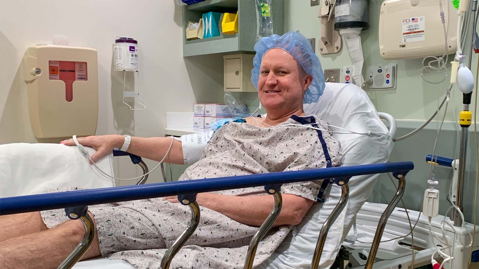 Scott in a hospital gown smiling while sitting on a bed, preparing for medical treatment with monitoring devices attached.