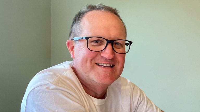 Scott smiling amd sitting against a green wall, wearing a white shirt.